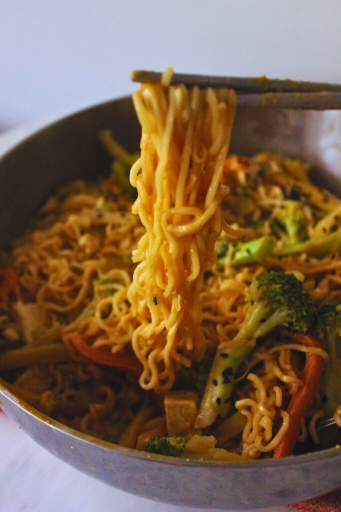 Image of chopsticks holding peanut noodles over a pan with peanut noodles and veggies