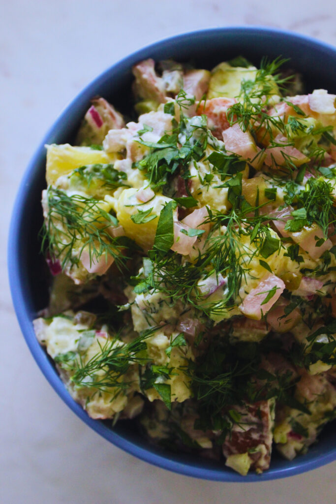 overhead image of a light blue bowl with potato salad topped with fresh herbs on a marble table top