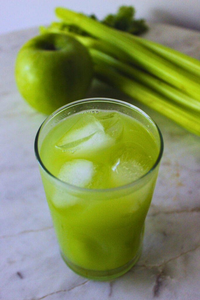 served glass of celery apple juice with ice on a marble table top with fresh green apple and celery stalk in the background