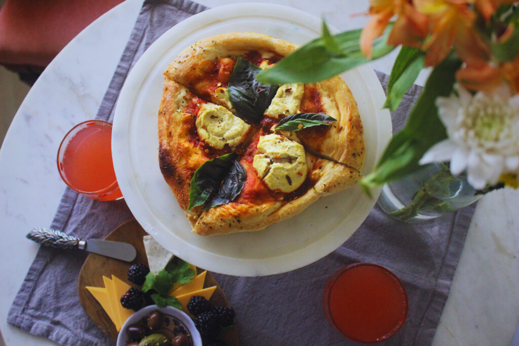 overhead photo of homemade vegan pizza with cashew ricotta and fresh basil with a side of cheese, olives and fruit and two glasses with a pink drink