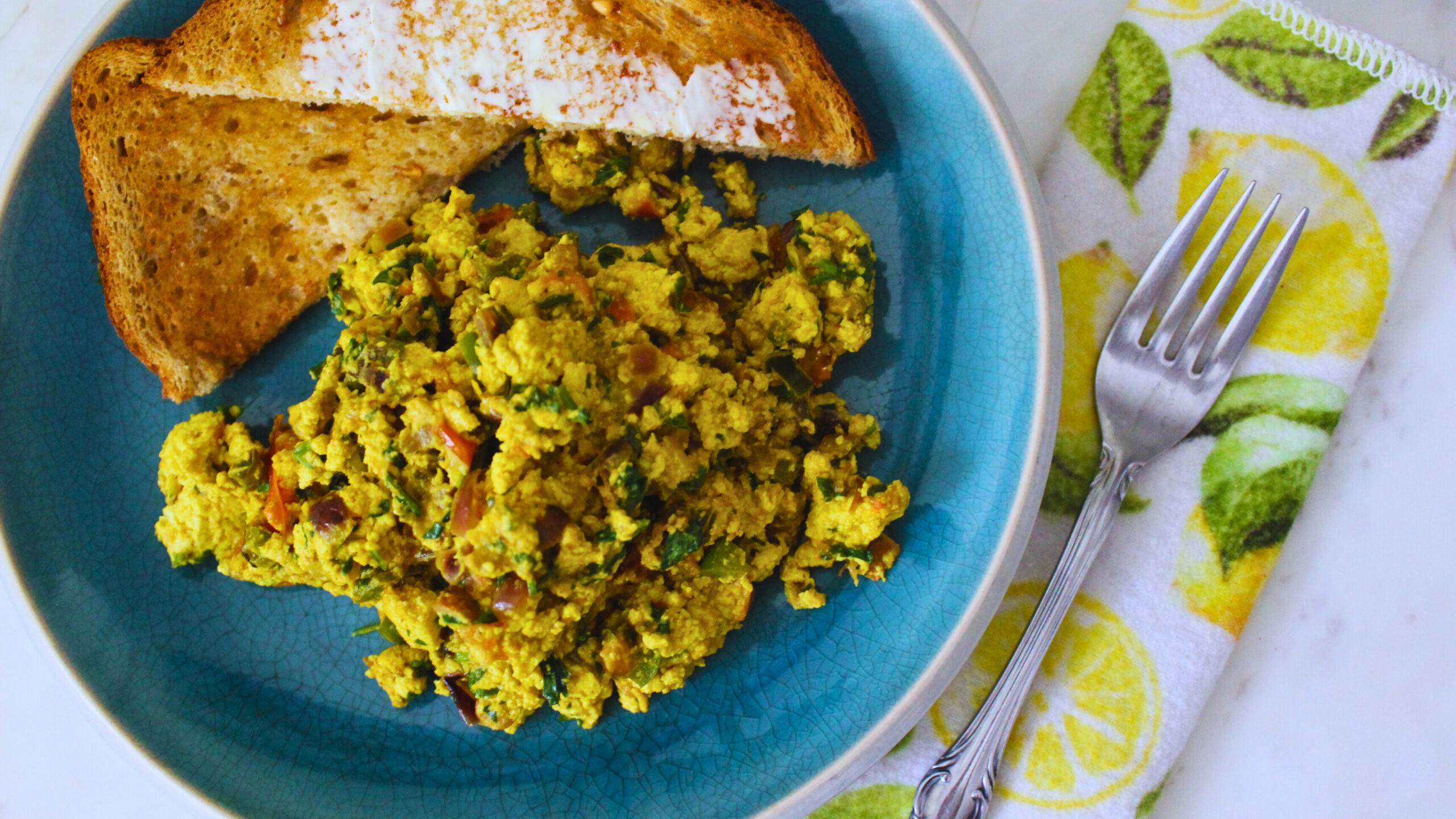 Image from above of tofu scramble and buttered toast on a plate, a fork sits on a napkin with lemon print next to it.