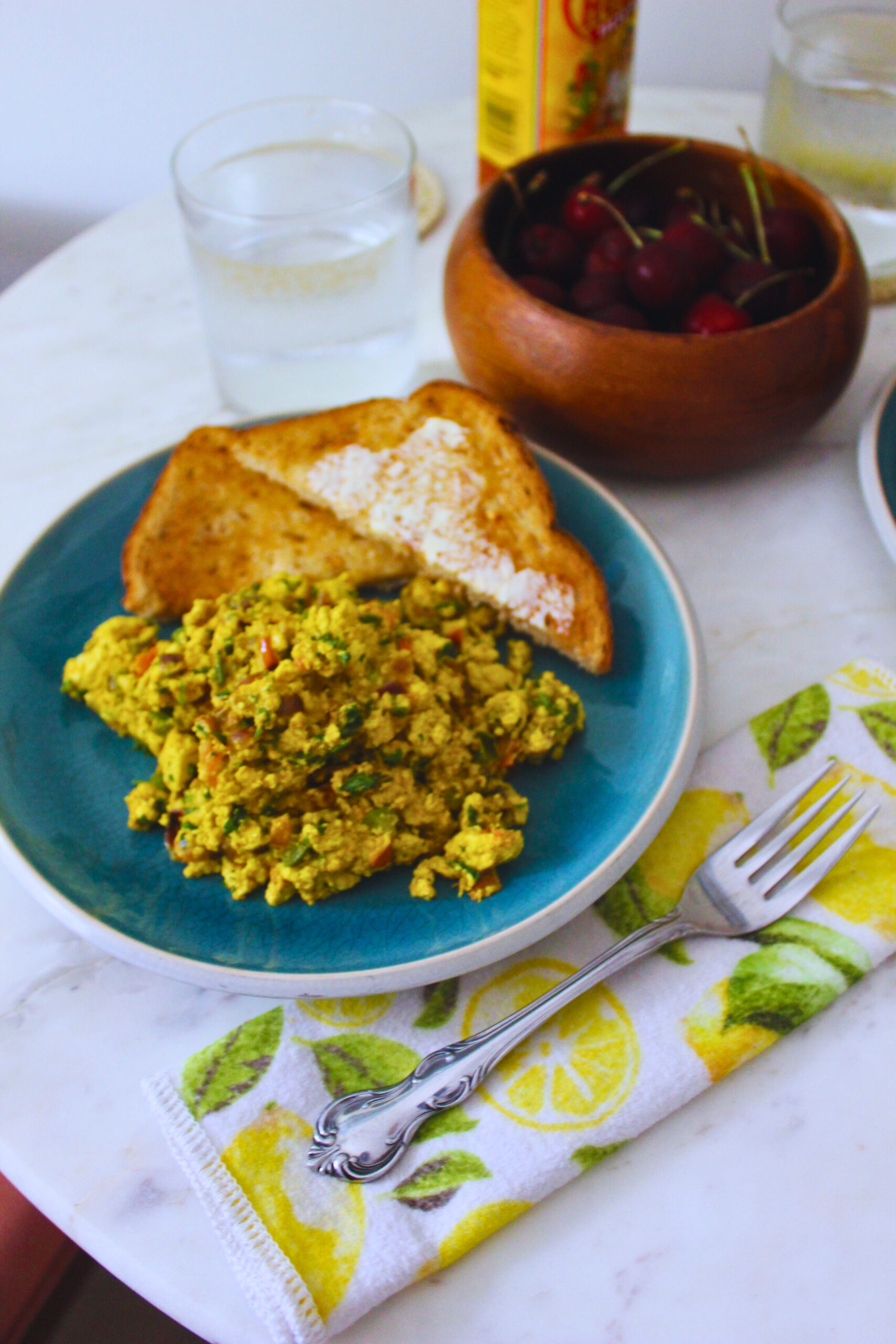 Image of tofu scramble and buttered toast on a plate, with a bowl of cherries, cholula, and a two glasses of water in the background.