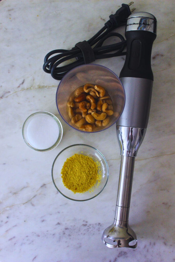 Overhead image of immersion blender, soaked cashews in a container, nutritional yeast in on a glass dish, and a salt container.