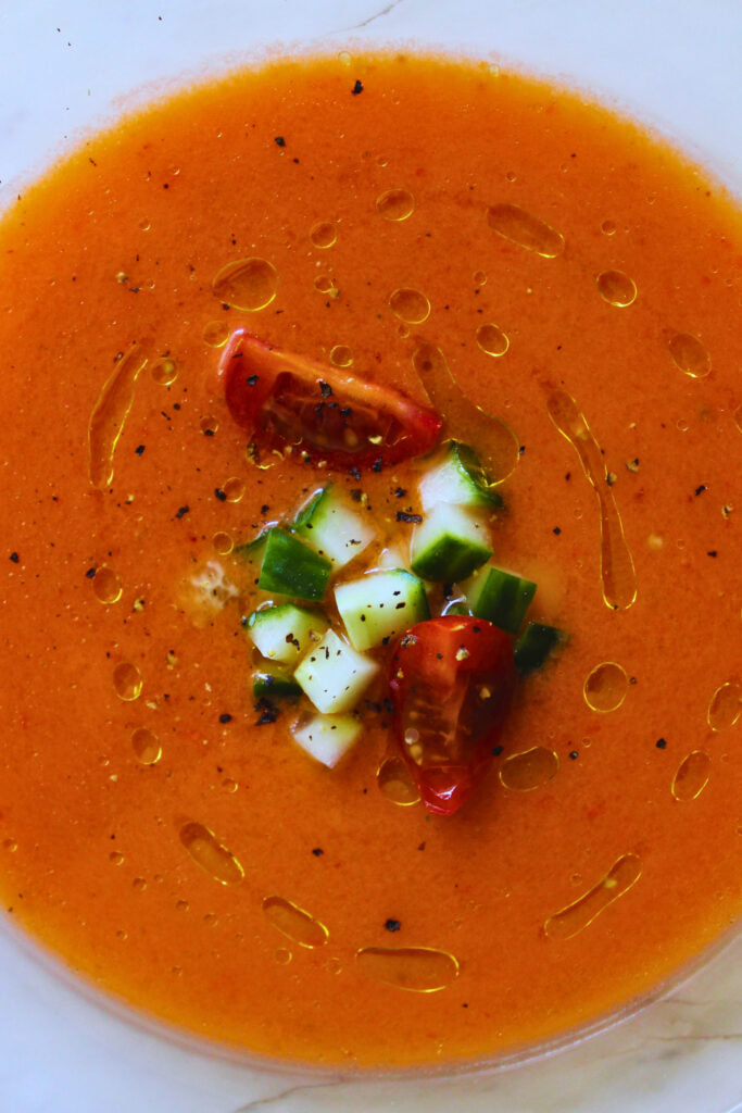 overhead image of gazpacho plated and topped with chopped cucumber, cherry tomatoes, and a drizzle of extra virgin olive oil
