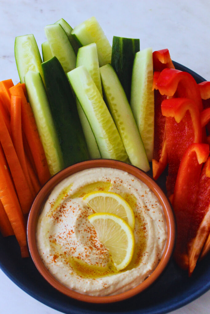 Overhead image of hummus surrounded by batons of carrots, cucumber, and red pepper