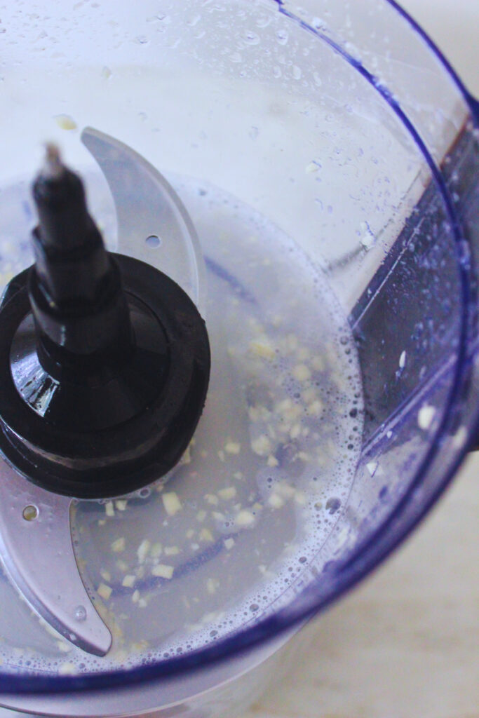 Overhead image of garlic resting in lemon juice in a food processor for a more mild garlic bite in the finished hummus