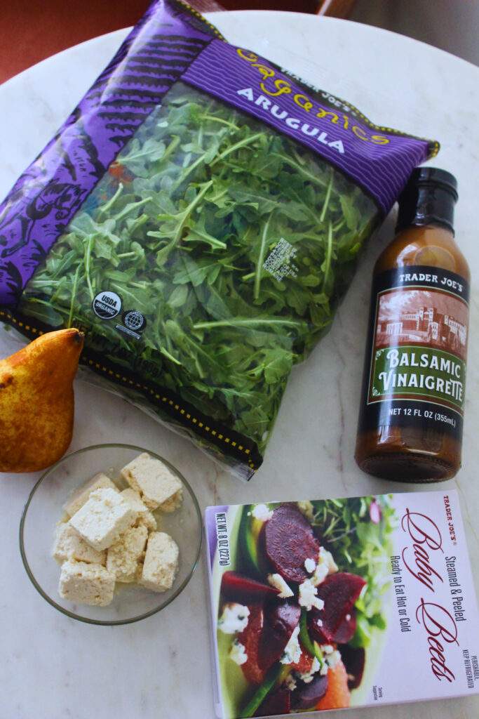 image of ingredients. bag of arugula, cooked beets, balsamic vinaigrette bottle, one pear, and a side of tofu feta.