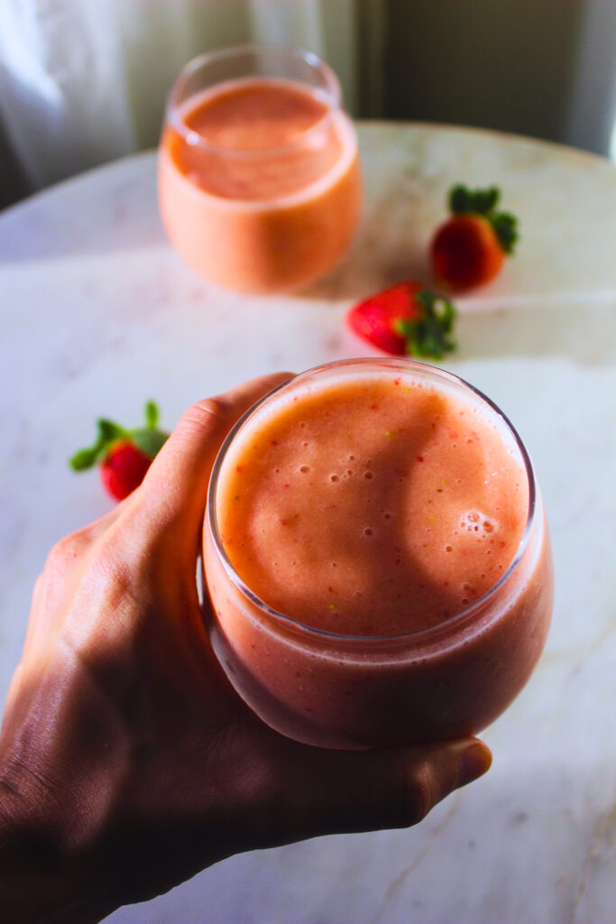 image of a hand holding a glass of the strawberry pineapple smoothie over a marble table in the early morning sun