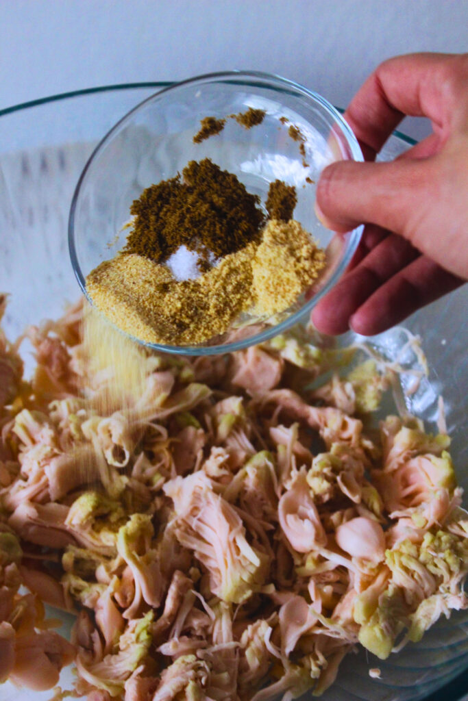 image of a hand tossing in spices on the shredded jackfruit in a bowl