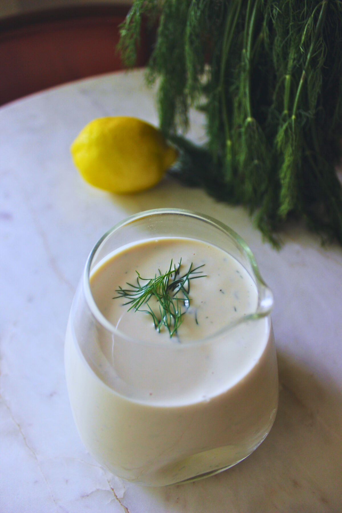 Image of a small glass jar with homemade dill pickle dressing. In the background there is a fresh lemon and fresh dill