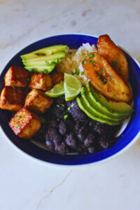 Bowl filled with rice, black beans, tofu, sweet plantains, avocado, topped with fresh lime and parsley