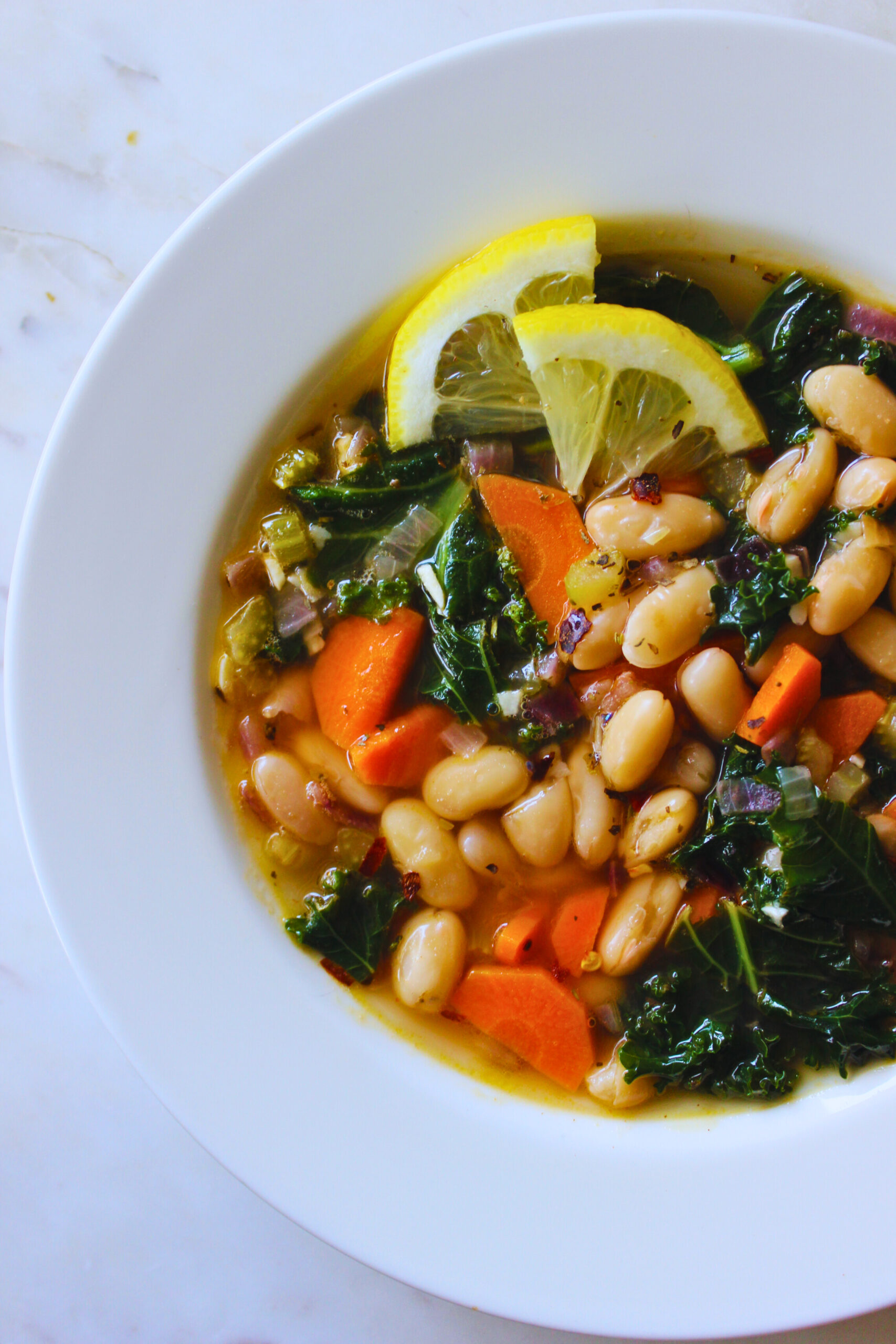 Prepared lemony kale and white bean soup. Served on a white plate topped with lemon wedge and red pepper flakes
