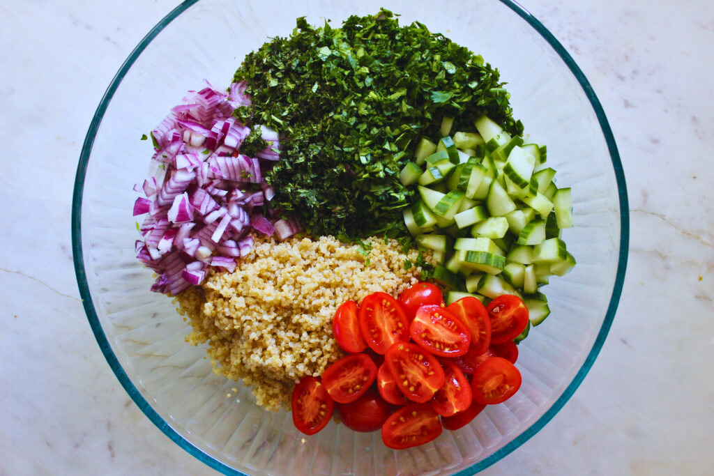 overhead image of large glass bowl filled with prepared ingredients for quinoa tabbouleh salad