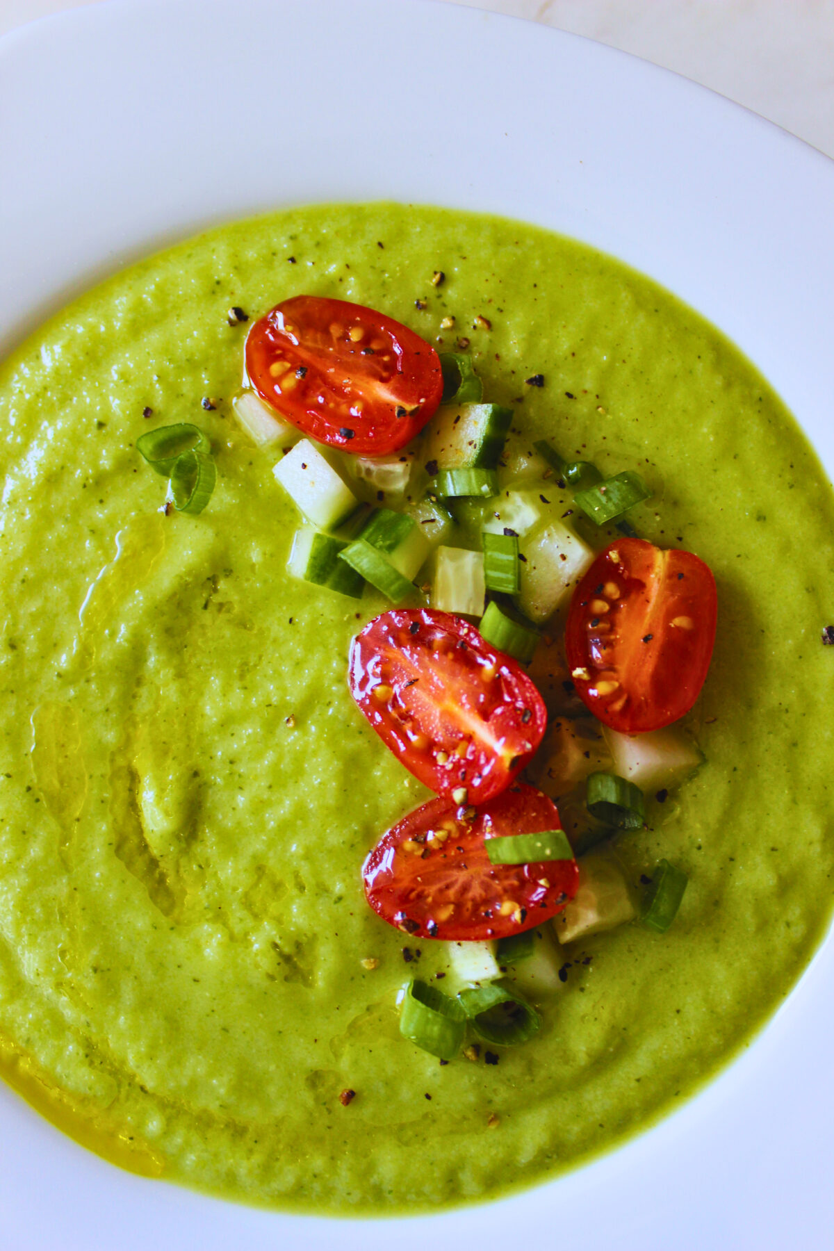 overhead image of green zucchini gazpacho topped with fresh tomatoes, cucumber and green onion.